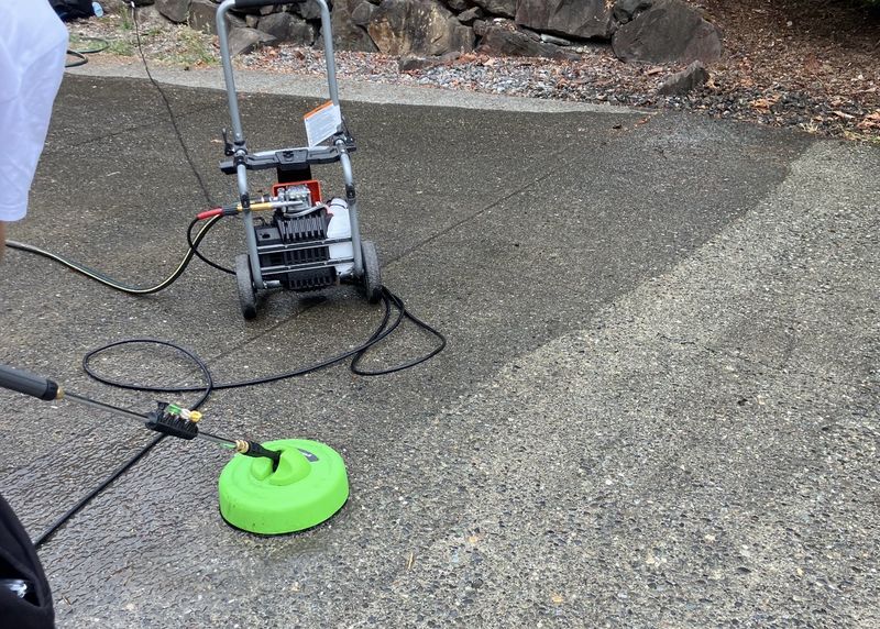 A pressure washer and green surface-cleaner attachment set up mid-job on a Sammamish driveway, with a rockery and landscaping behind.